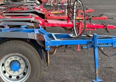 A row of several trailer dollies in blue and red parked closely together on a gravel lot. The foreground features a blue dolly with its landing leg extended, showing detailed views of the heavy-duty tires, silver wheel rims, and hydraulic hose connections.