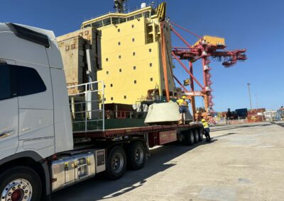 A white Volvo semi-truck stationary at a port while a large yellow crane lowers a heavy concrete structure onto its red trailer. Two workers in high-visibility vests are supervising the loading process next to a large yellow ship.