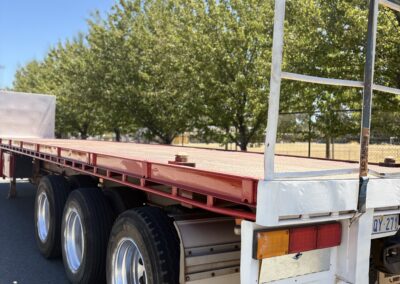 Rear side view of a triple-axle flatbed trailer with red steel deck and Energy Hire branding, parked on a paved area under clear blue skies.