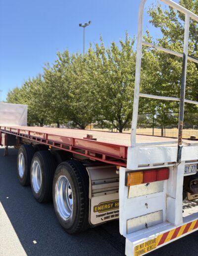 Rear side view of a triple-axle flatbed trailer with red steel deck and Energy Hire branding, parked on a paved area under clear blue skies.
