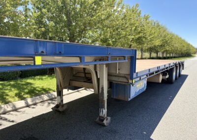 A low-angle shot showing the front landing gear and underside of a blue step deck trailer. The trailer has a tan-colored deck and a blue "Energy Hire" storage box mounted to the frame. A row of green trees lines the road in the background.
