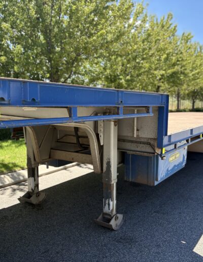 A low-angle shot showing the front landing gear and underside of a blue step deck trailer. The trailer has a tan-colored deck and a blue "Energy Hire" storage box mounted to the frame. A row of green trees lines the road in the background.