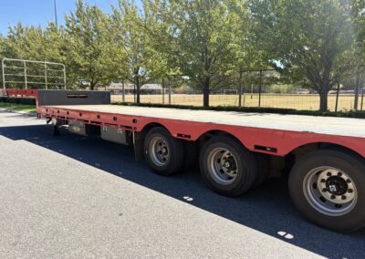 A wide-angle side view of a long, red drop deck semi-trailer parked on an asphalt road. The trailer features a silver checkered plate deck, three axles at the rear, and a white safety railing at the front. Green trees and a chain-link fence line the background under a clear blue sky.