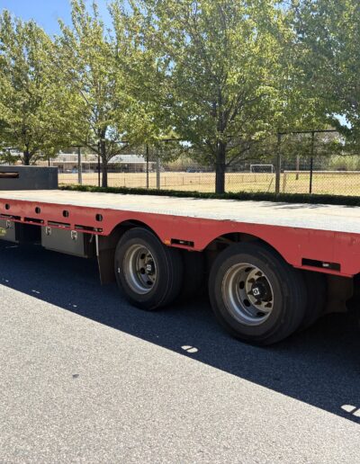 A wide-angle side view of a long, red drop deck semi-trailer parked on an asphalt road. The trailer features a silver checkered plate deck, three axles at the rear, and a white safety railing at the front. Green trees and a chain-link fence line the background under a clear blue sky.