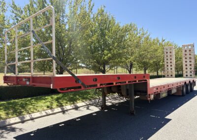 A red drop deck trailer shown from a side-rear perspective parked on a road. It features a white safety headboard railing at the front and two tall, white loading ramps standing vertically at the rear.