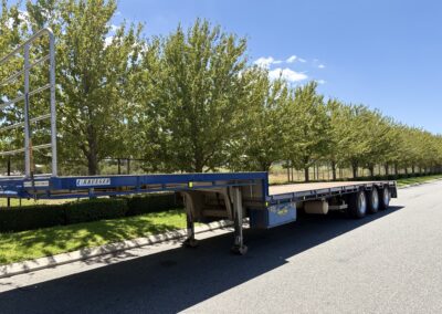 A blue Krueger brand drop deck trailer parked along a tree-lined street. The trailer has a silver headboard railing, a tan-colored deck, and blue storage boxes mounted to the chassis between the landing gear and rear axles.