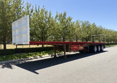 A profile view of a red flat top trailer parked on a sunny road. It features a large, solid white headboard at the front and a smaller silver safety railing at the rear, with red "Energy Hire" toolboxes visible under the deck.