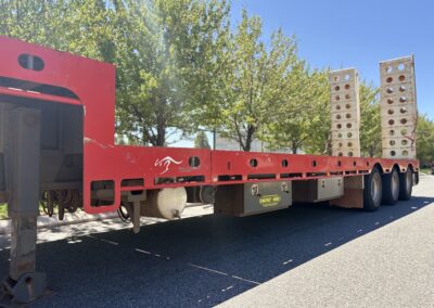 A low-angle profile of a red drop deck trailer parked on a suburban street. The trailer features a white kangaroo decal, grey "Energy Hire" toolboxes, and two upright white perforated loading ramps at the rear.