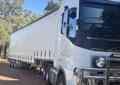 A white Volvo Globetrotter semi-truck with a large silver bull bar parked on a red dirt track, pulling a long white curtainsider trailer through a wooded area.