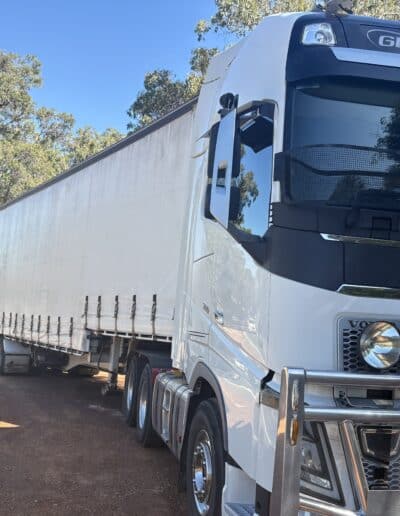 A white Volvo Globetrotter semi-truck with a large silver bull bar parked on a red dirt track, pulling a long white curtainsider trailer through a wooded area.